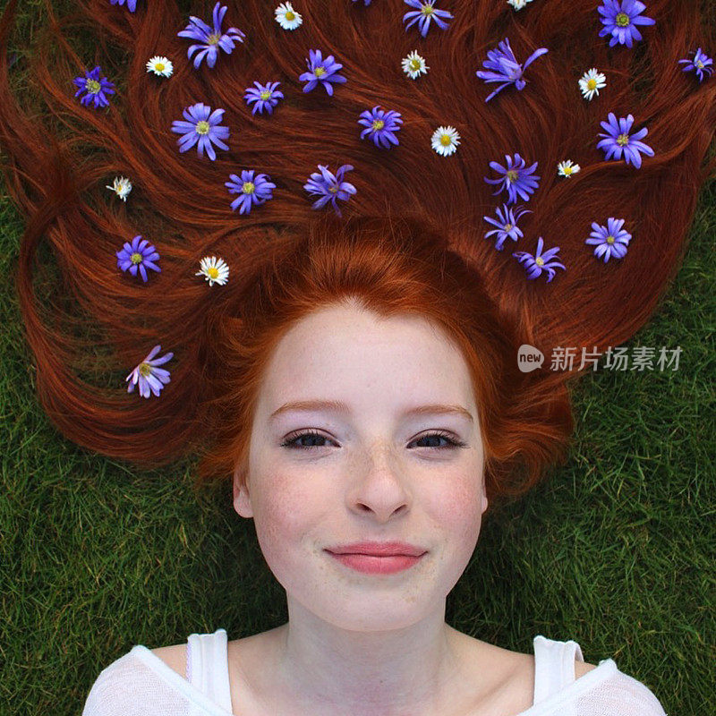 14 / 15岁的红发少女躺在草地上，皮肤苍白，脸上长着雀斑，头发散布在她的头上，点缀着白色的雏菊和紫色的花朵